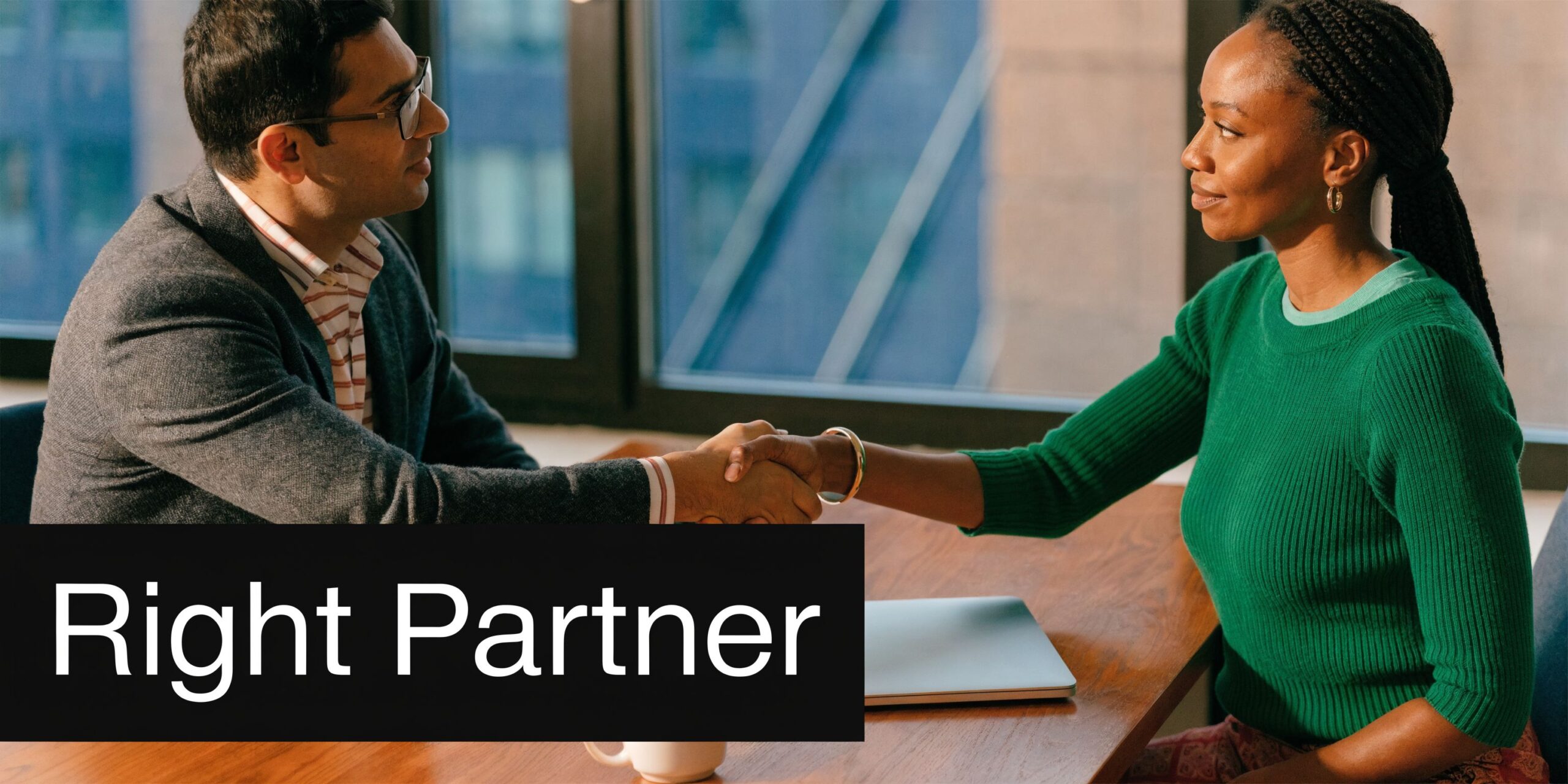 A professional man and woman sitting at a desk shaking hands in a bright modern office setting.