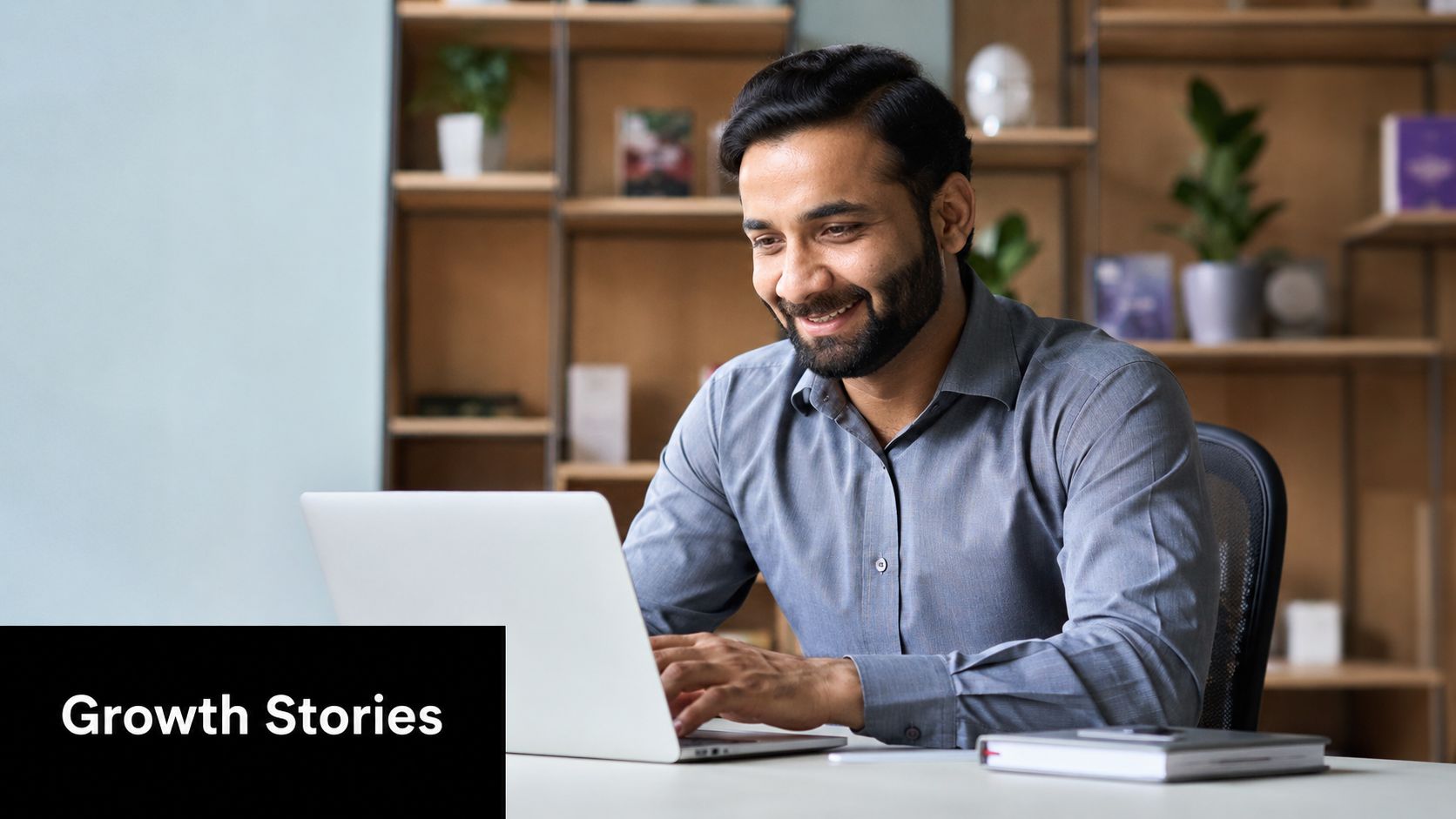 A professional man with a beard smiling while working on his laptop in a modern office.